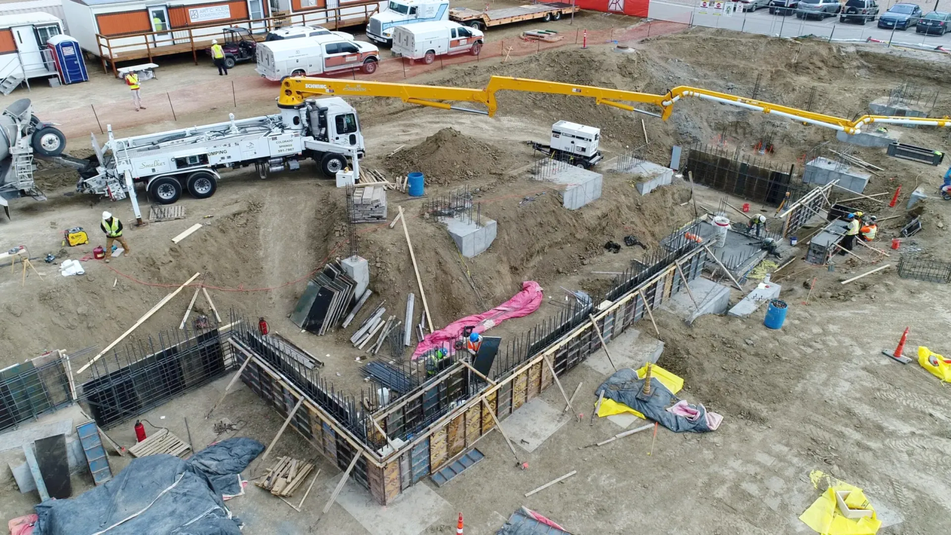 An aerial view of a construction site with workers, concrete forms, and a large pump truck on a dirt excavation lot.