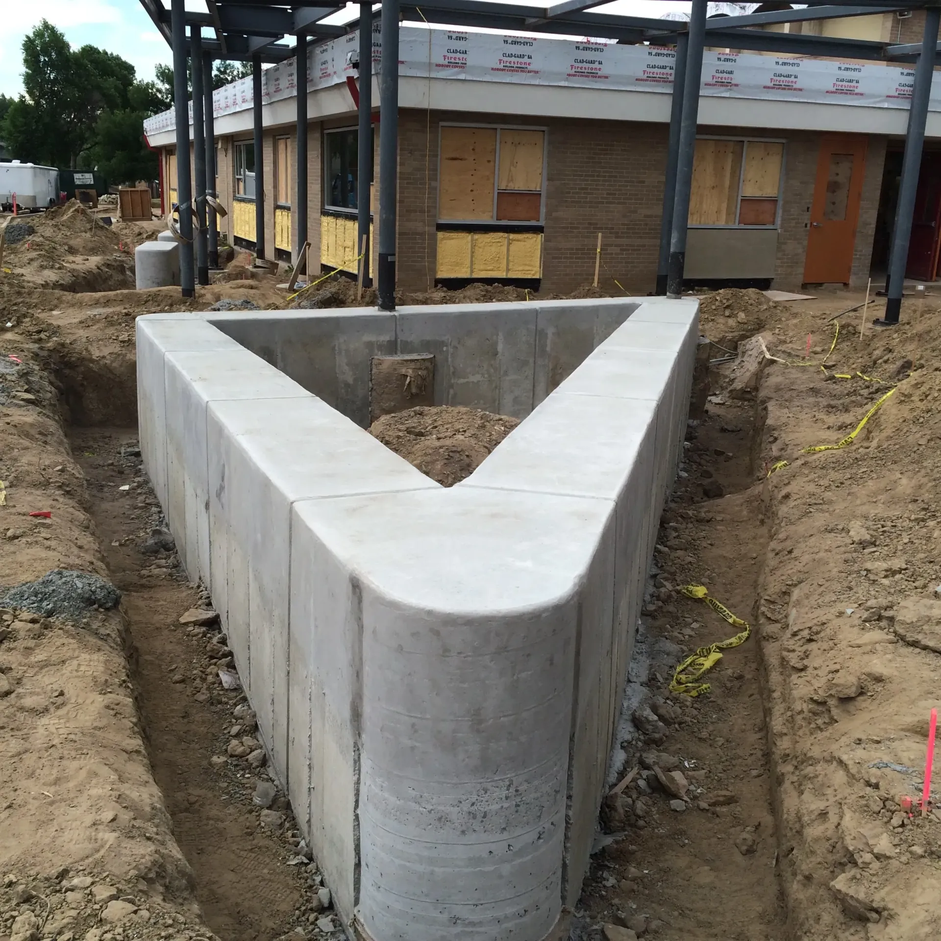 A V-shaped concrete retaining wall structure in a dirt construction site with a building in the background.