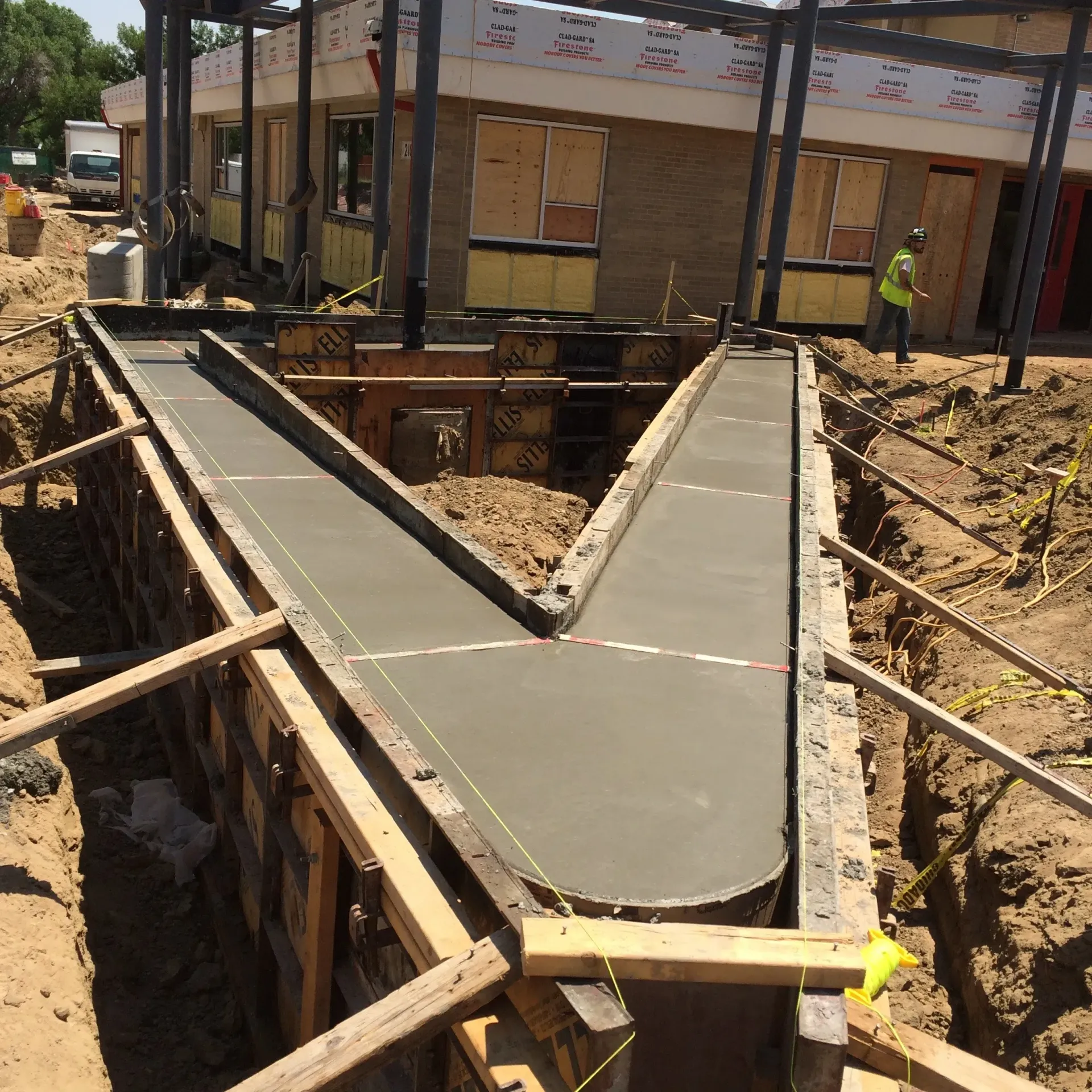 Freshly poured V-shaped concrete footings in a construction trench in front of a building under renovation.
