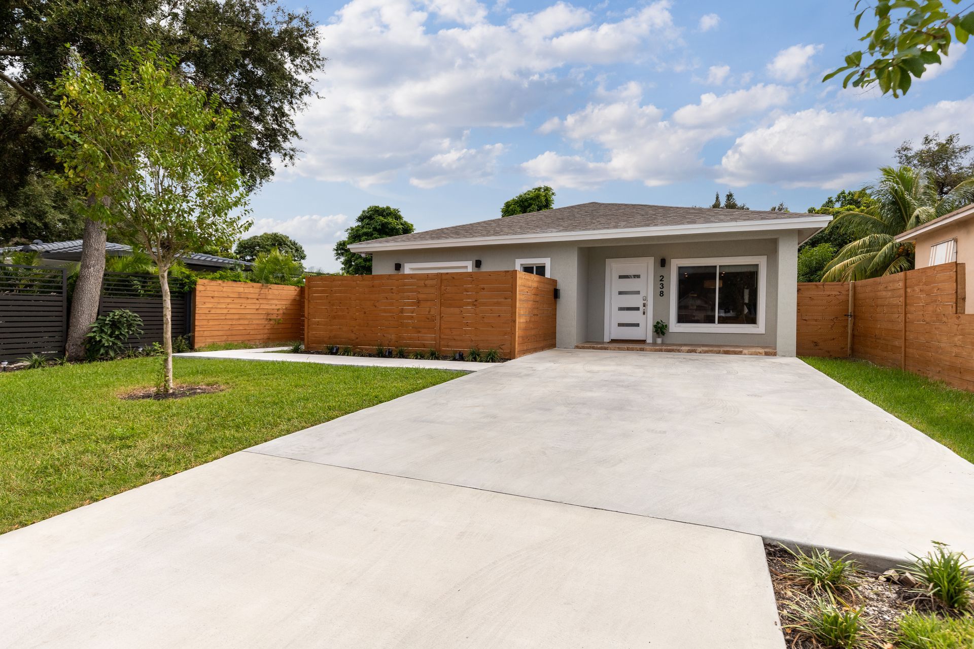 Single-story house with gray stucco, a white door, a concrete driveway, and a wooden slat fence under a partly cloudy sky.