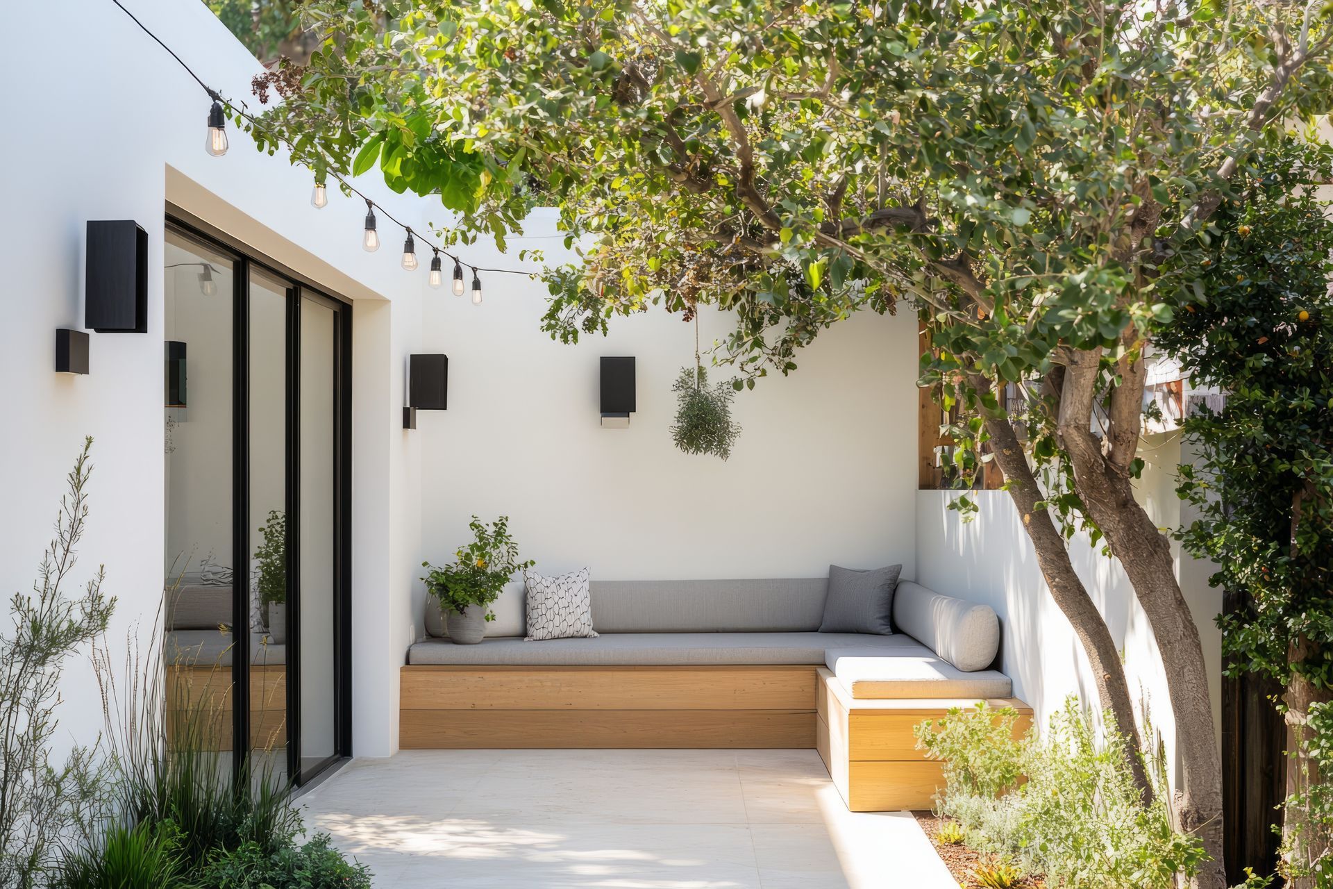 An L-shaped wooden built-in bench with grey cushions sits in a white-walled outdoor courtyard under a tree.