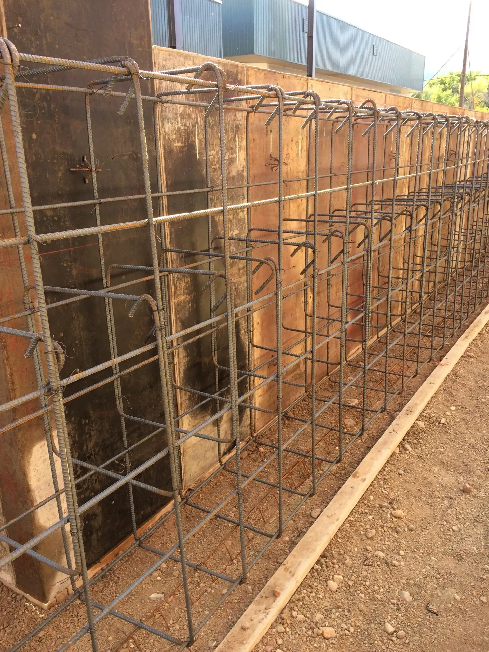 A construction site showing a series of rectangular rebar cages positioned in a row against a dark concrete wall.