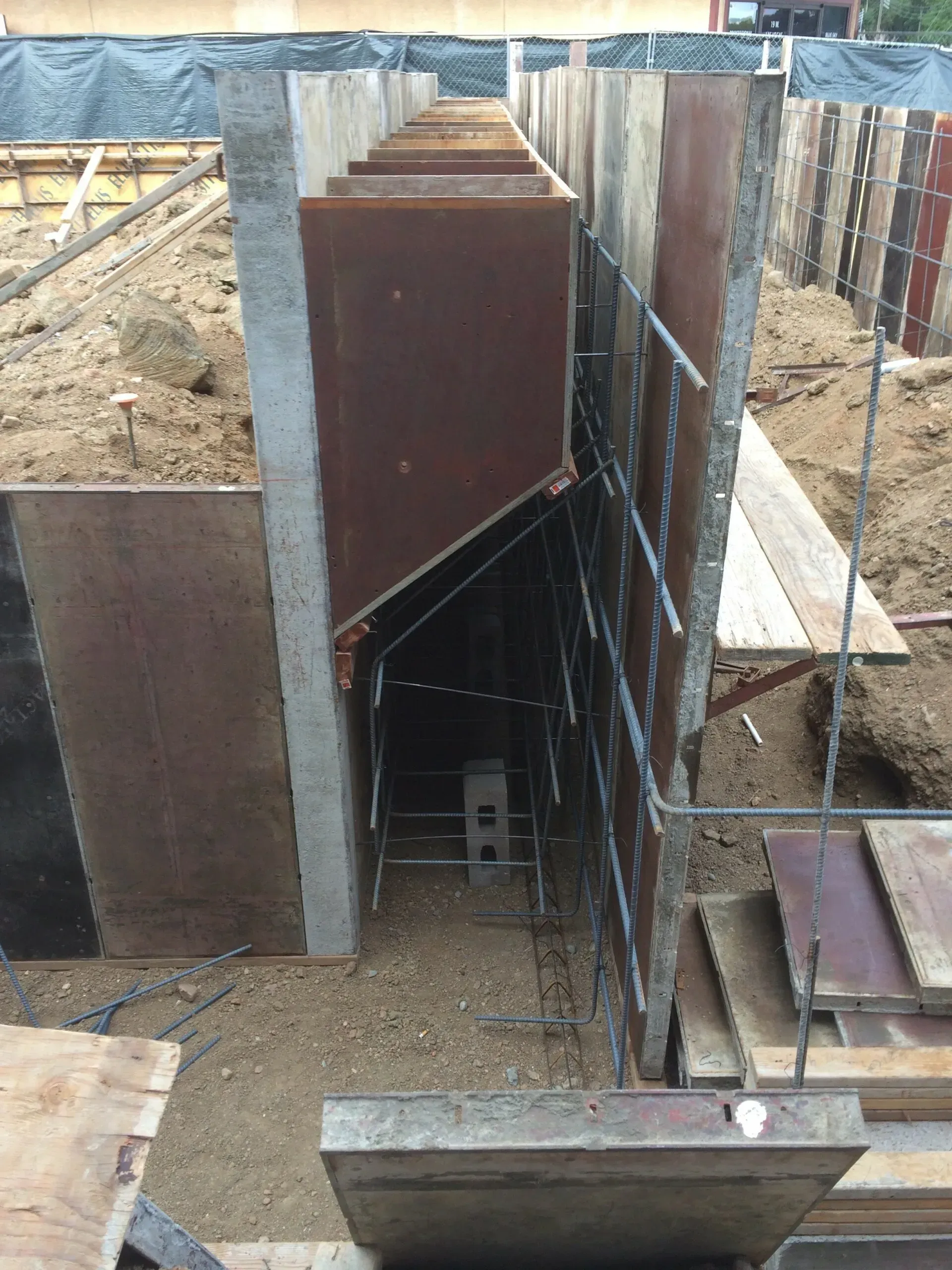 A construction site showing concrete forms set up for a foundation wall, with exposed steel rebar cages in the trench.