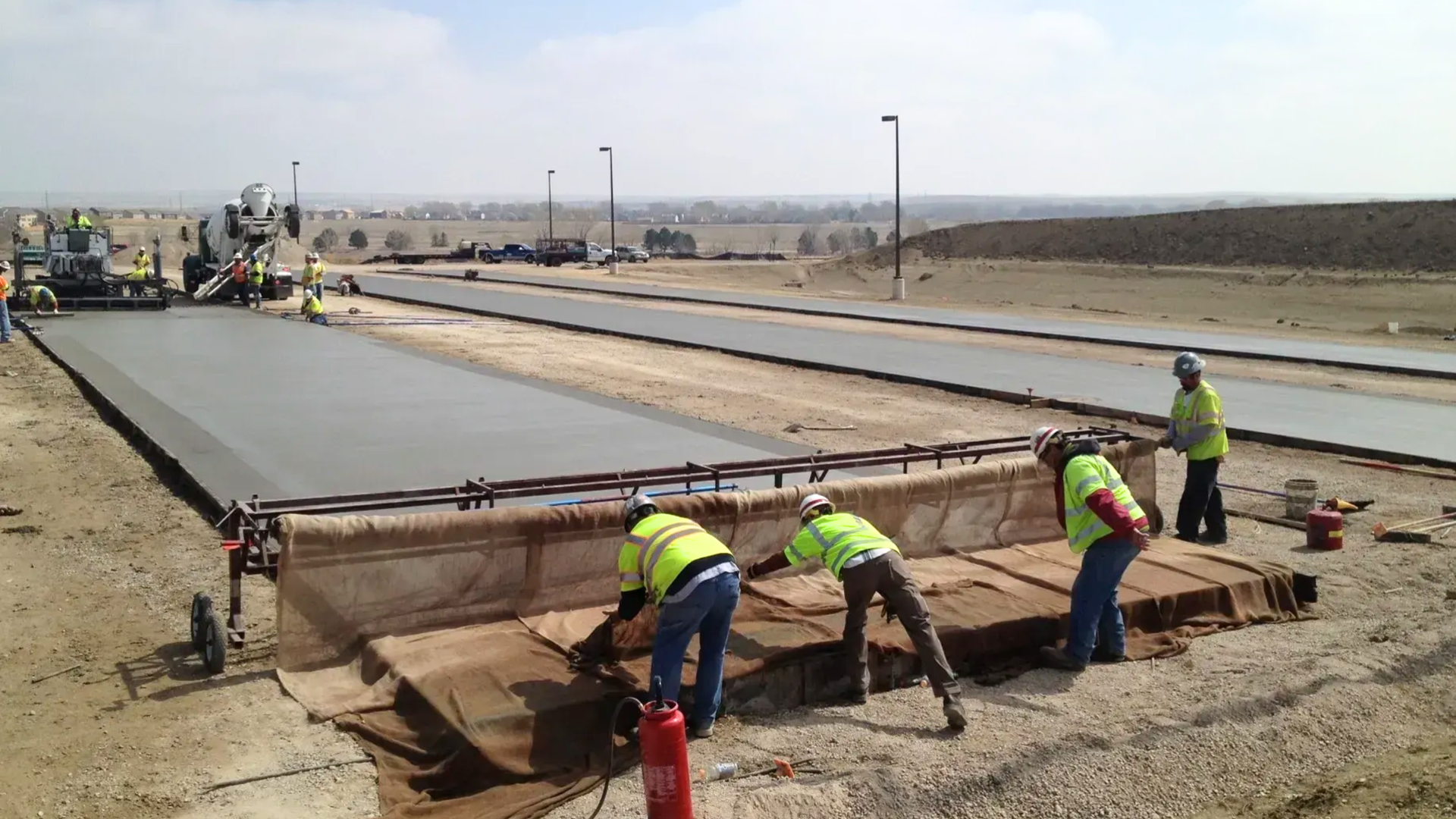 Construction workers in high-visibility vests use a burlap-covered screed to finish a freshly poured concrete roadway.
