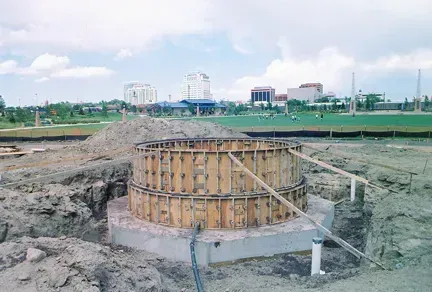 A circular concrete construction form sits in an excavated foundation at a park-like site with buildings in the background.