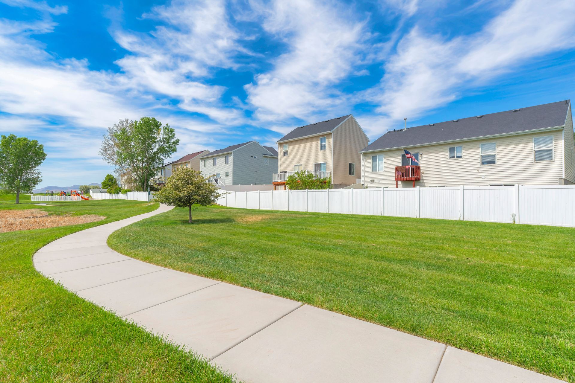 A concrete path curves through a grassy park toward several houses with white fences on a sunny, blue-sky day.