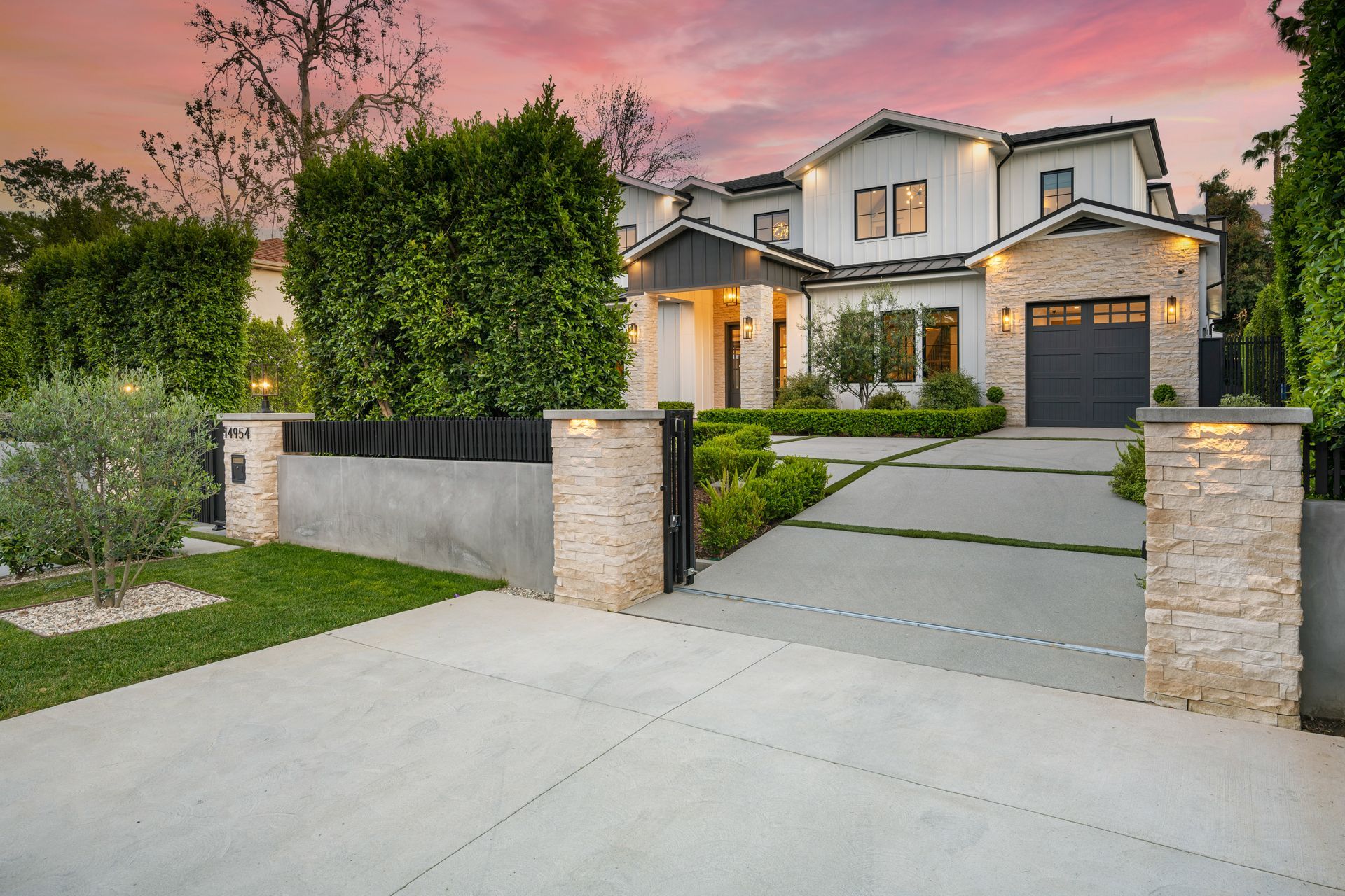 Modern two-story house with a stone facade, a concrete driveway, and lush landscaping under a sunset sky.