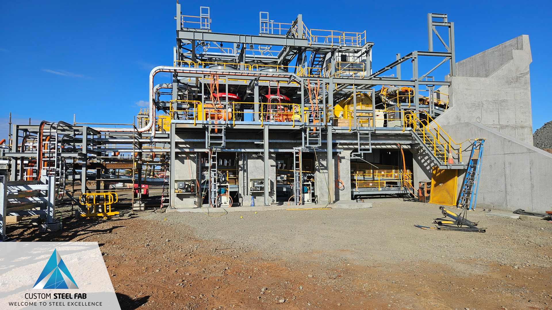 Industrial processing plant with metal scaffolding, pipes, and gravel yard under a clear blue sky