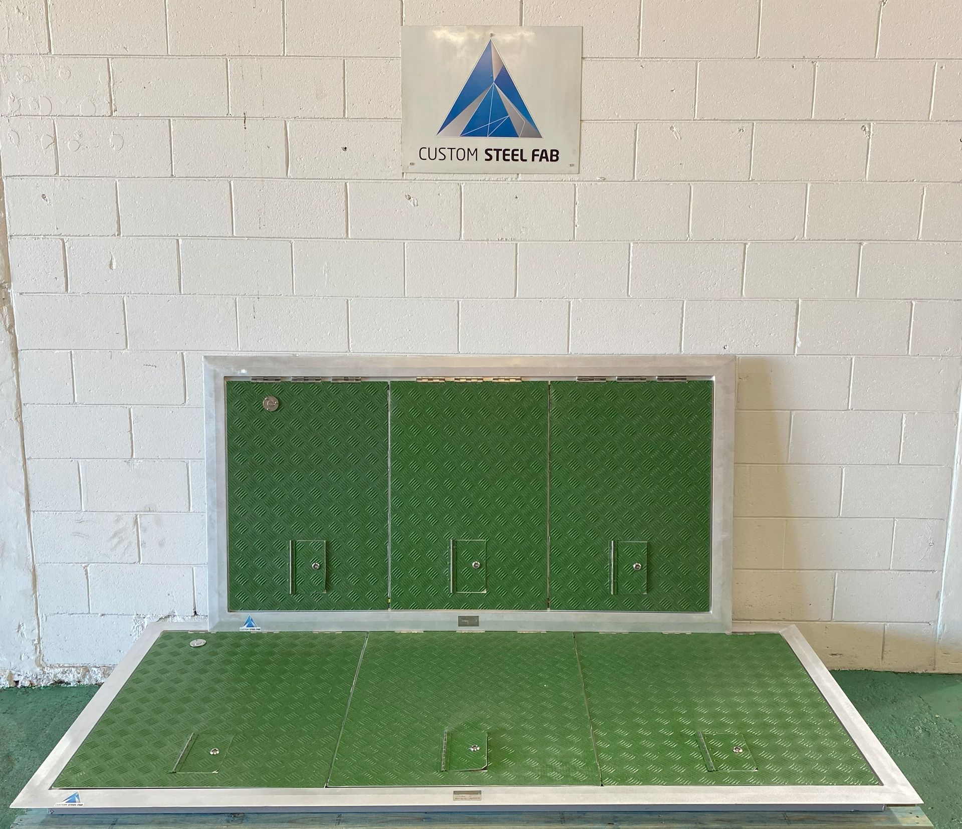 Green Metal Utility Covers on a White Brick Wall and Floor Beneath a Company Sign — Custom Steel Fab in Bohle, QLD
