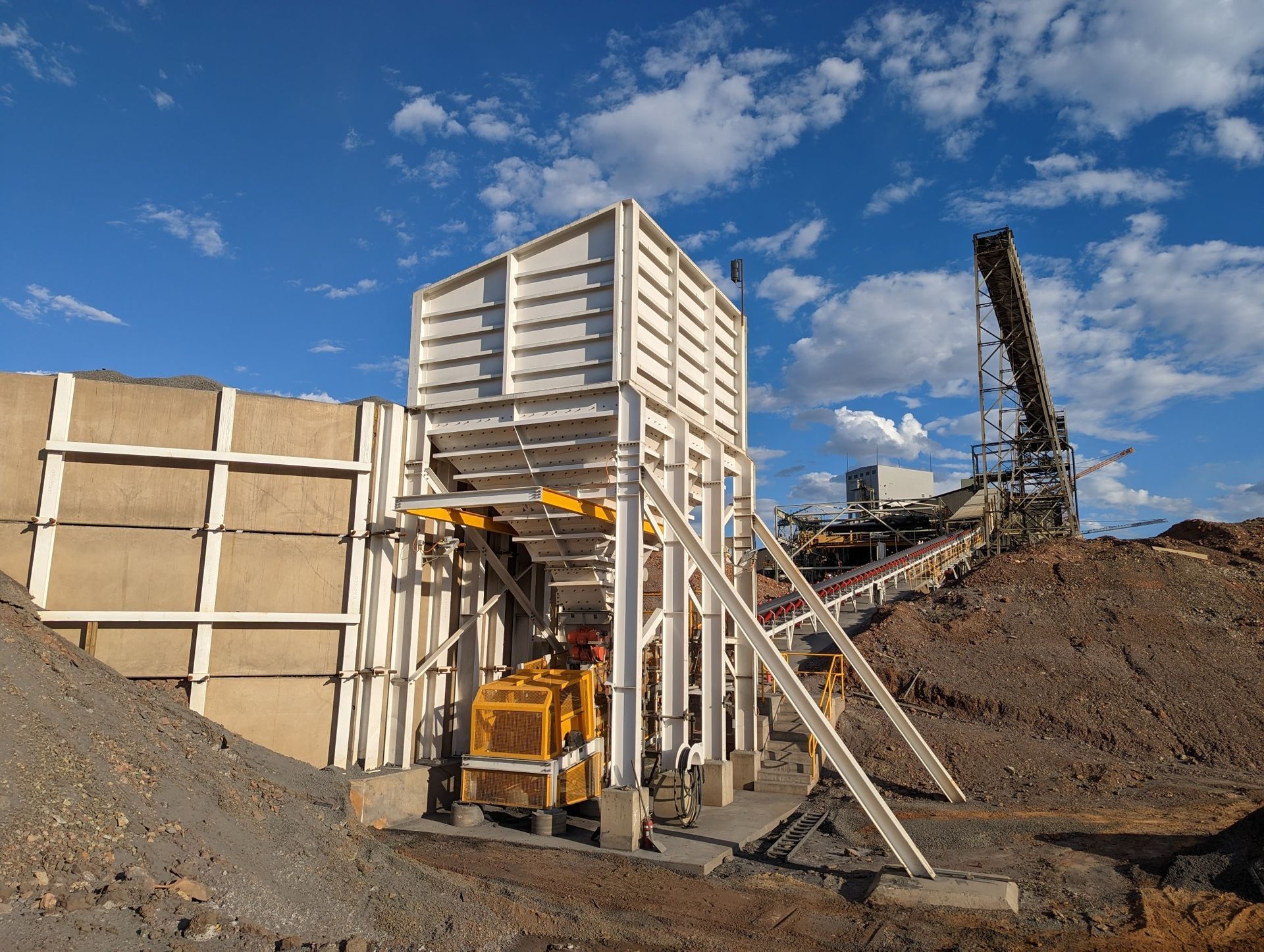 Construction site with a yellow excavator beside dirt piles and a partially built concrete structure under a blue sky