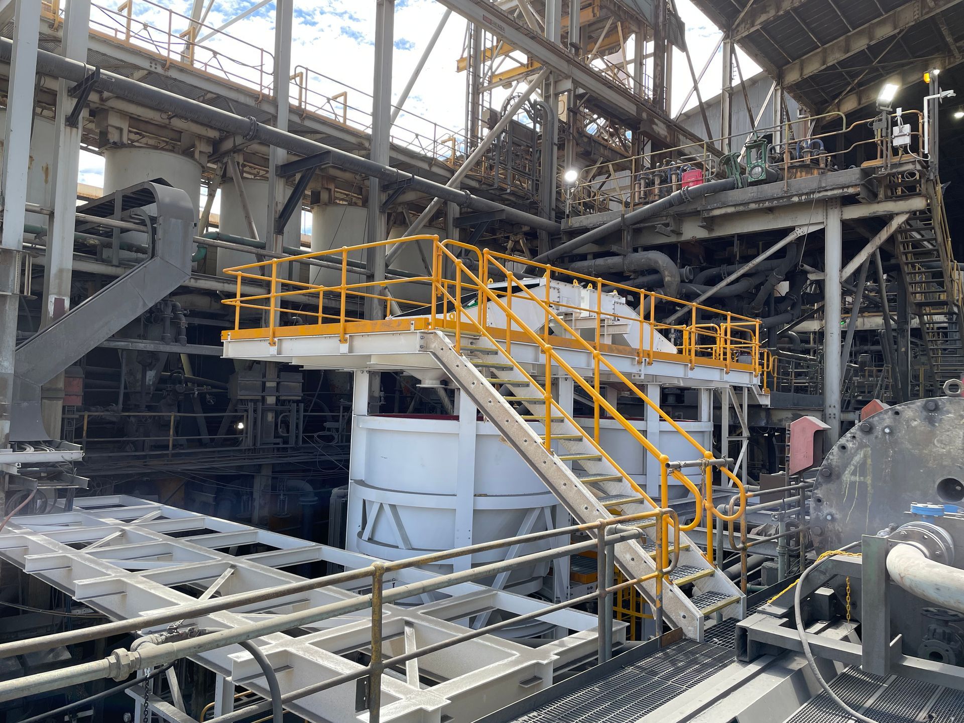 Industrial plant interior with yellow-railed staircase and large machinery under a high metal roof