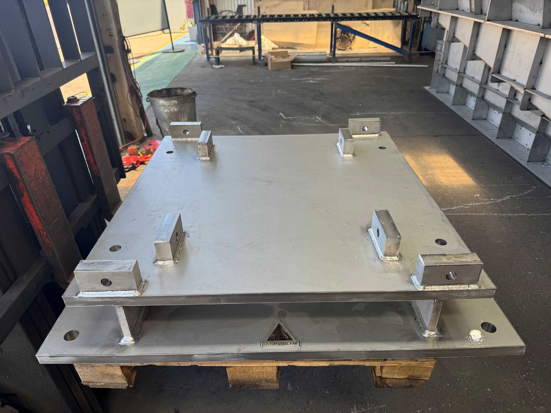 Metal Platform With Four Bolts on a Pallet in an Industrial Warehouse Aisle — Custom Steel Fab in Bohle, QLD