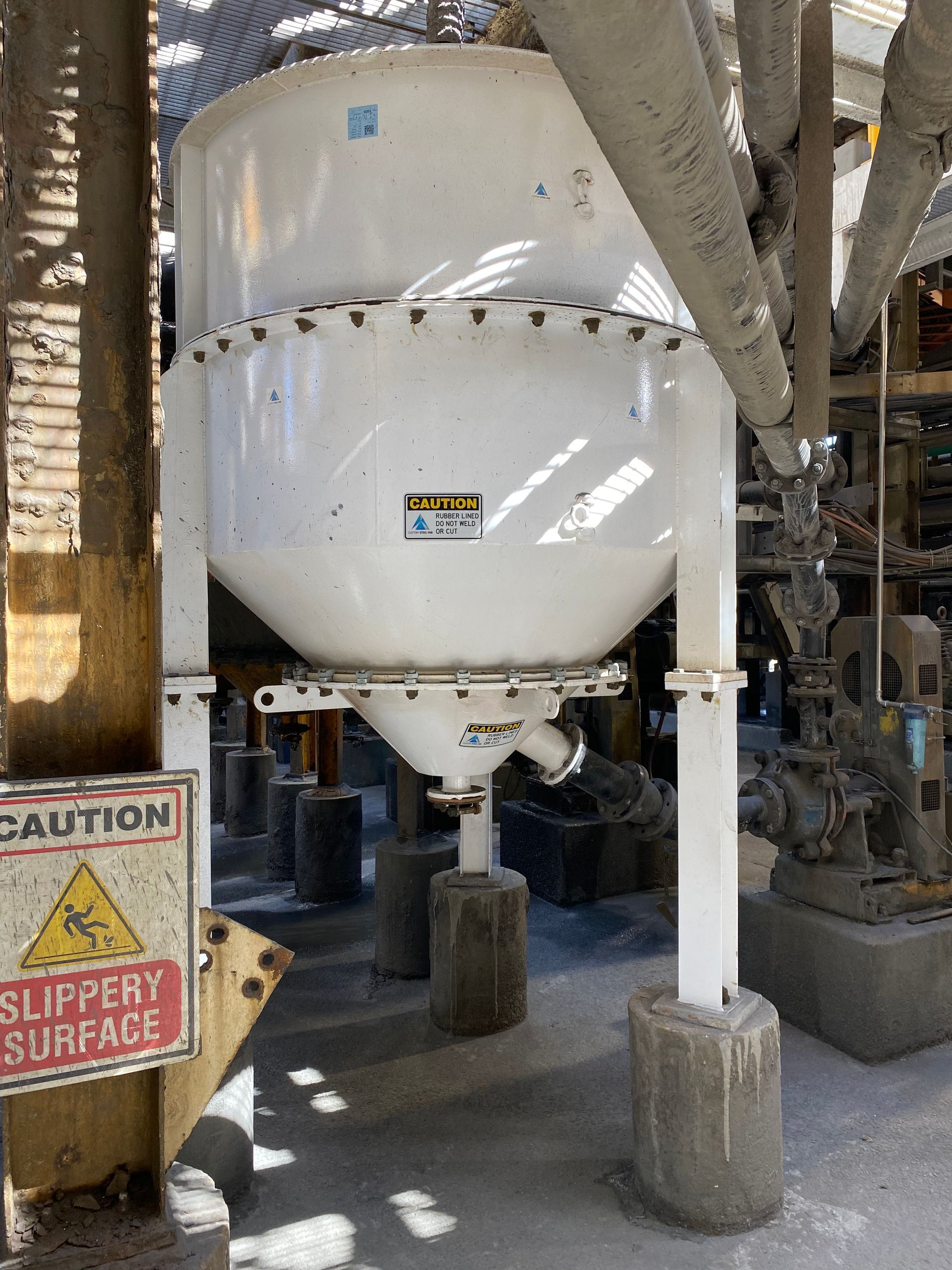 Large white industrial hopper tank on supports in a factory, with caution and slippery surface signs nearby