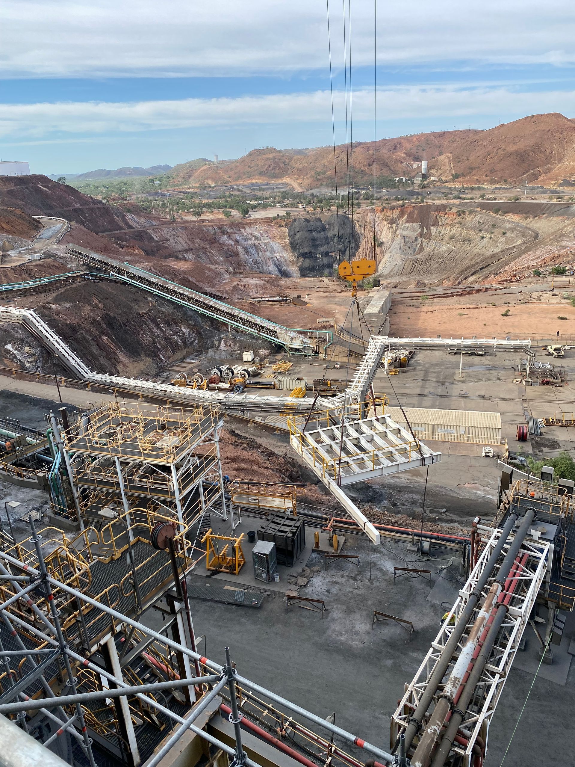 Open-pit mine with conveyor structures, a yellow excavator, and rocky hills under a clear sky