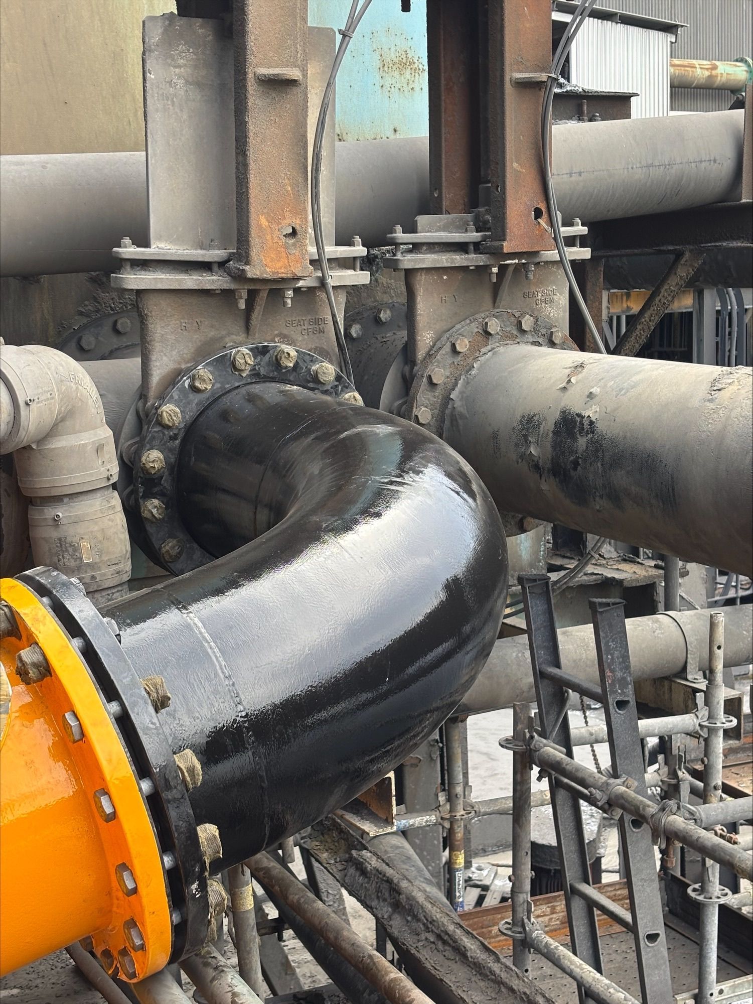 Large black industrial pipe with orange flange connected to rusty metal piping and valves.