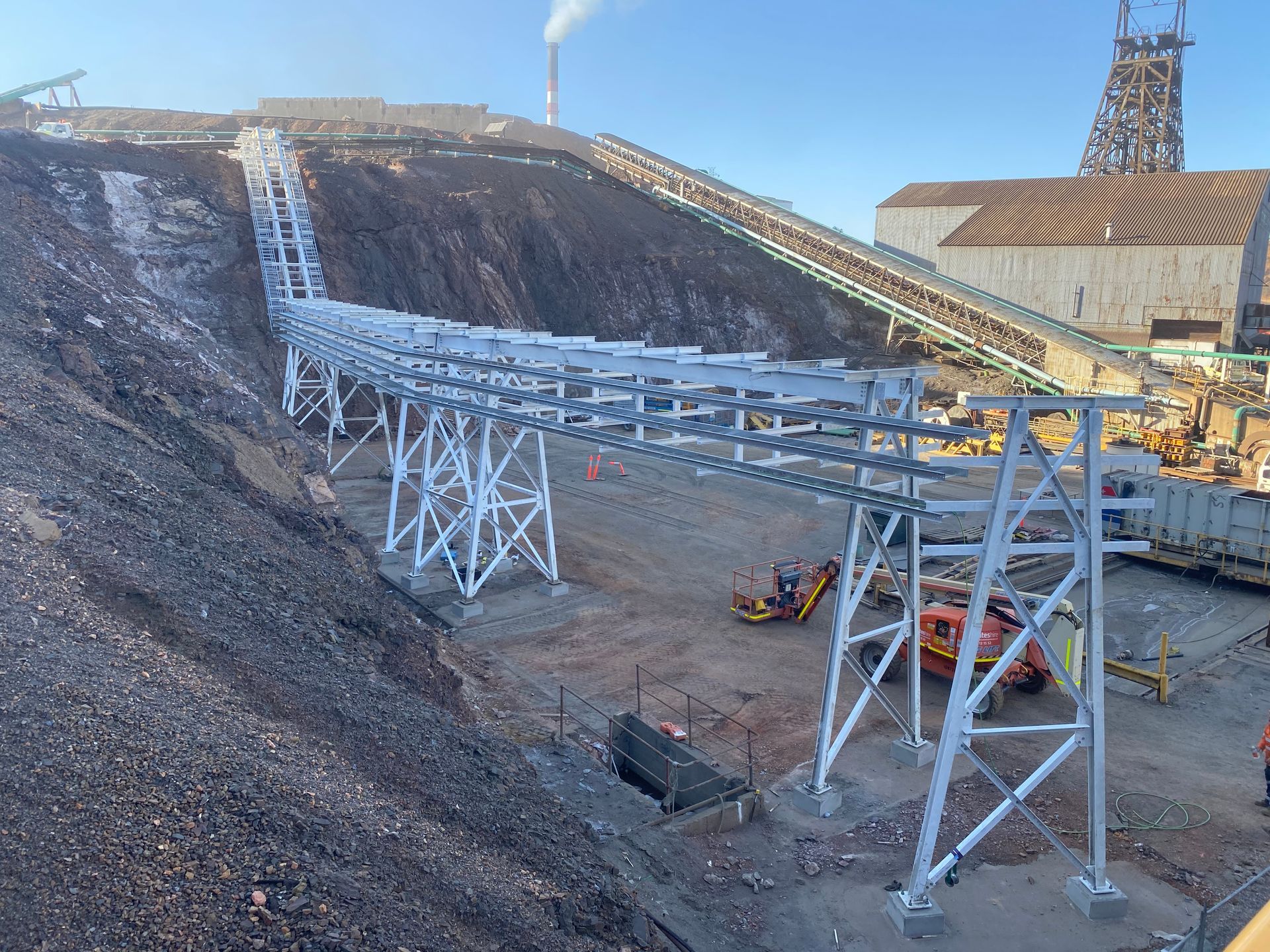 Conveyor structures at an open-pit mine with excavators and rocky slopes under a clear sky