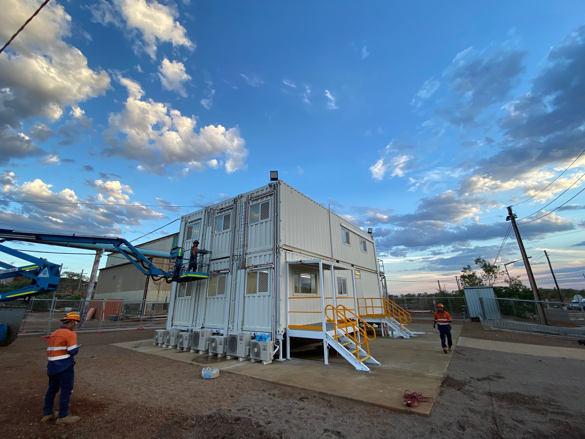 Industrial white container unit with stairs on a gravel lot under a blue cloudy sky, workers nearby