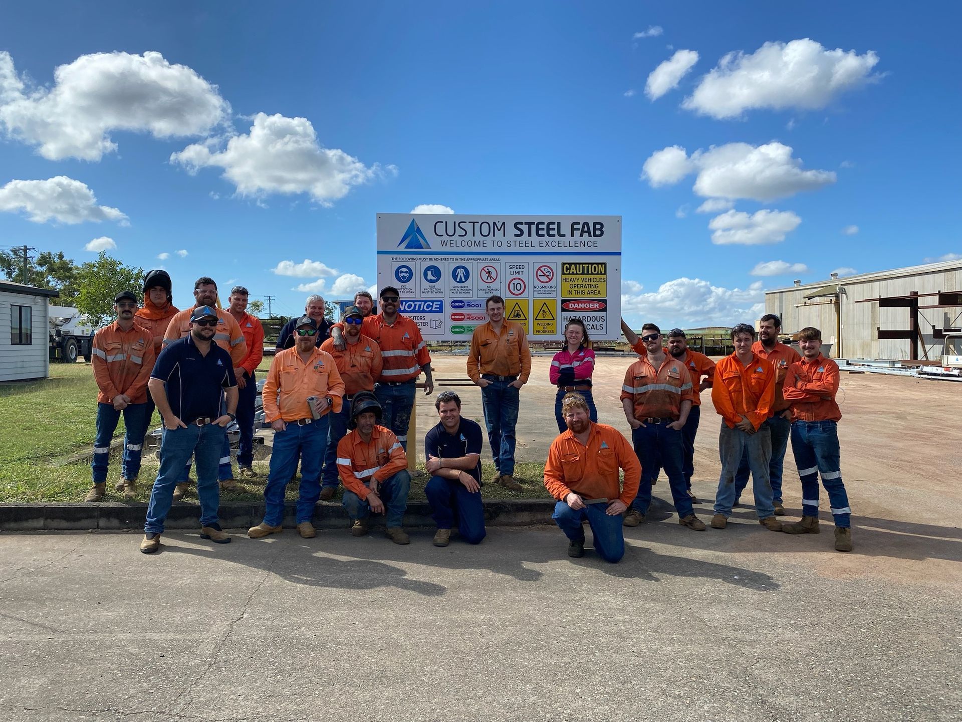 Group of Workers in Orange Safety Gear Posing Outdoors in Front of a Signboard — Custom Steel Fab in Bohle, QLD