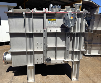 Large stainless steel industrial tank or hopper outdoors on metal legs, with pipes and warning label.