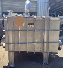 Large stainless steel industrial tank with pipes and caution sign outside a warehouse
