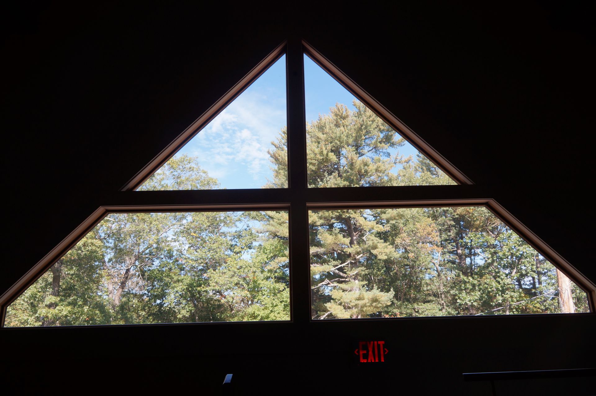 A triangular window with a cross-shaped frame looks out onto green trees against a bright blue sky.