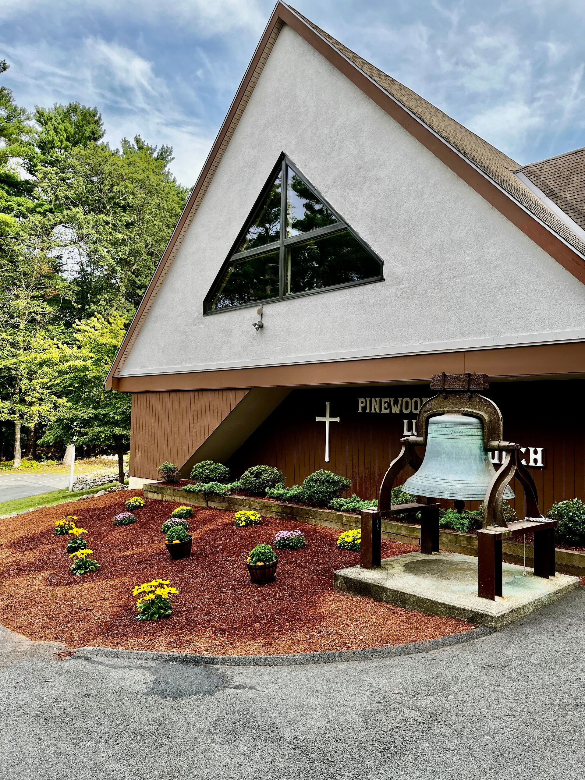 An A-frame church with a triangular window above a bell display in a mulched flower bed.