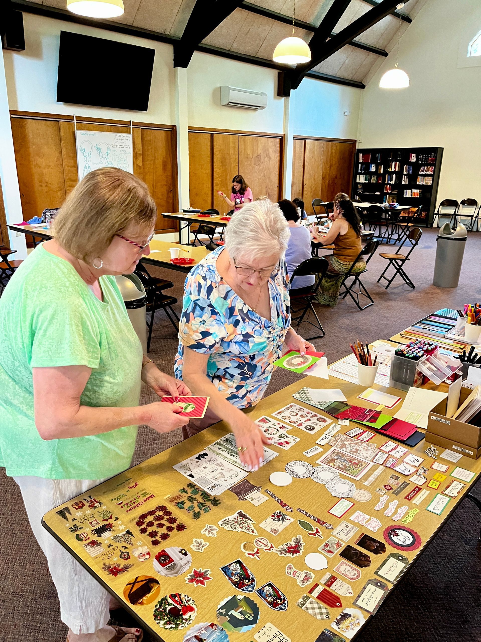 Two people standing at a table in a hall, arranging colorful stickers and paper crafts together.
