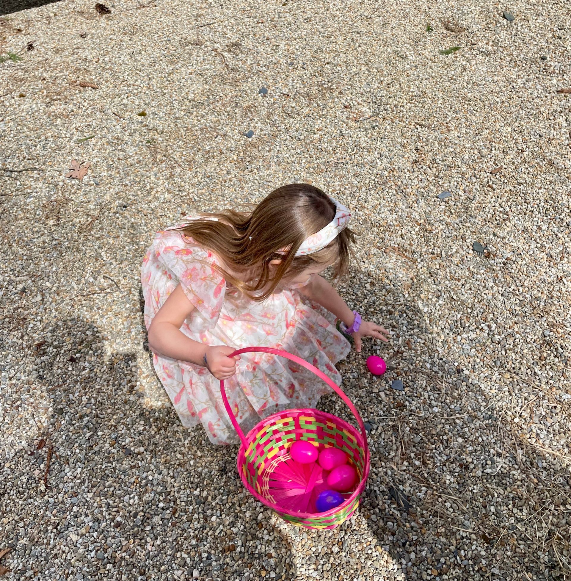 A person wearing a floral dress gathers colorful plastic eggs from a gravel ground into a woven basket.