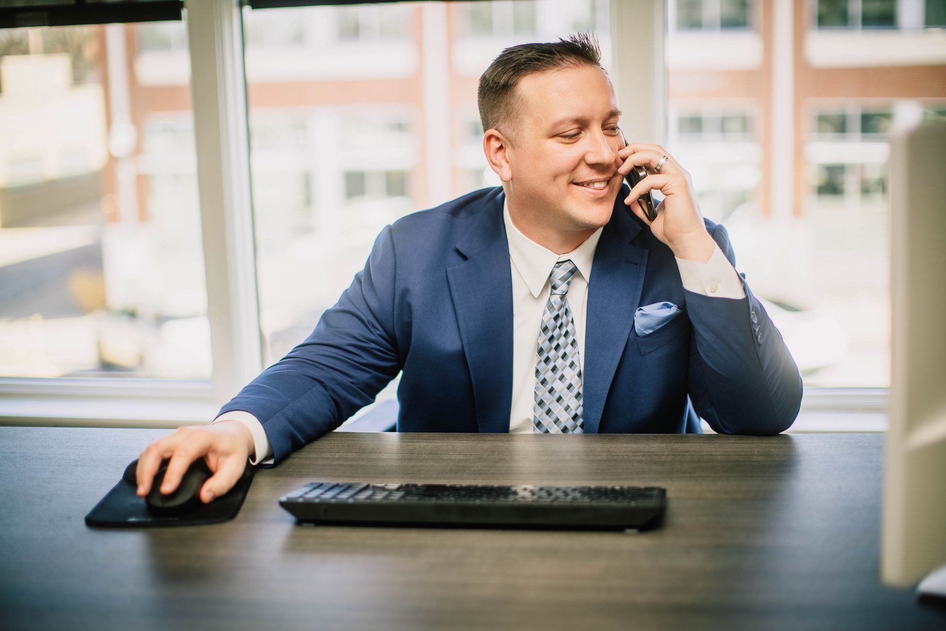 A man in a suit and tie is sitting at a desk talking on a cell phone.