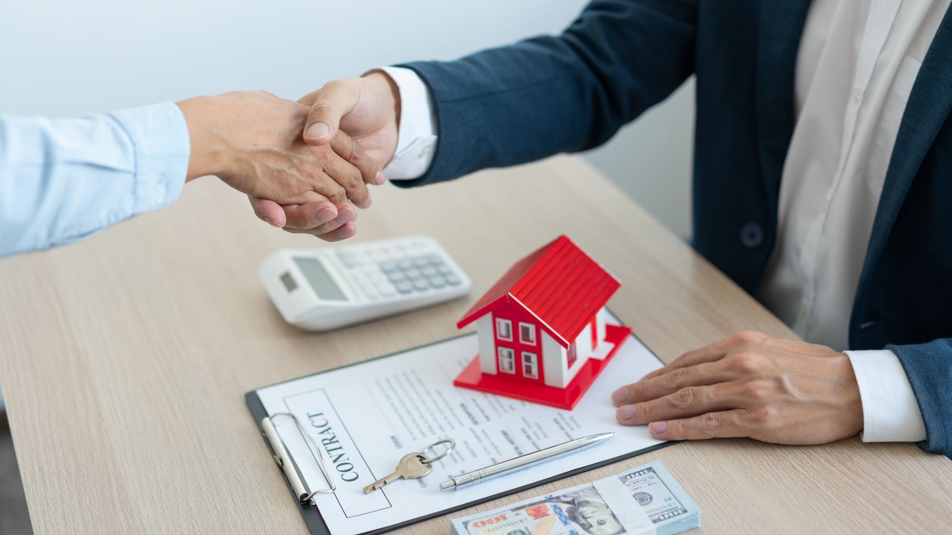 Two people shaking hands over a desk with a contract, house model, calculator, keys, and money.