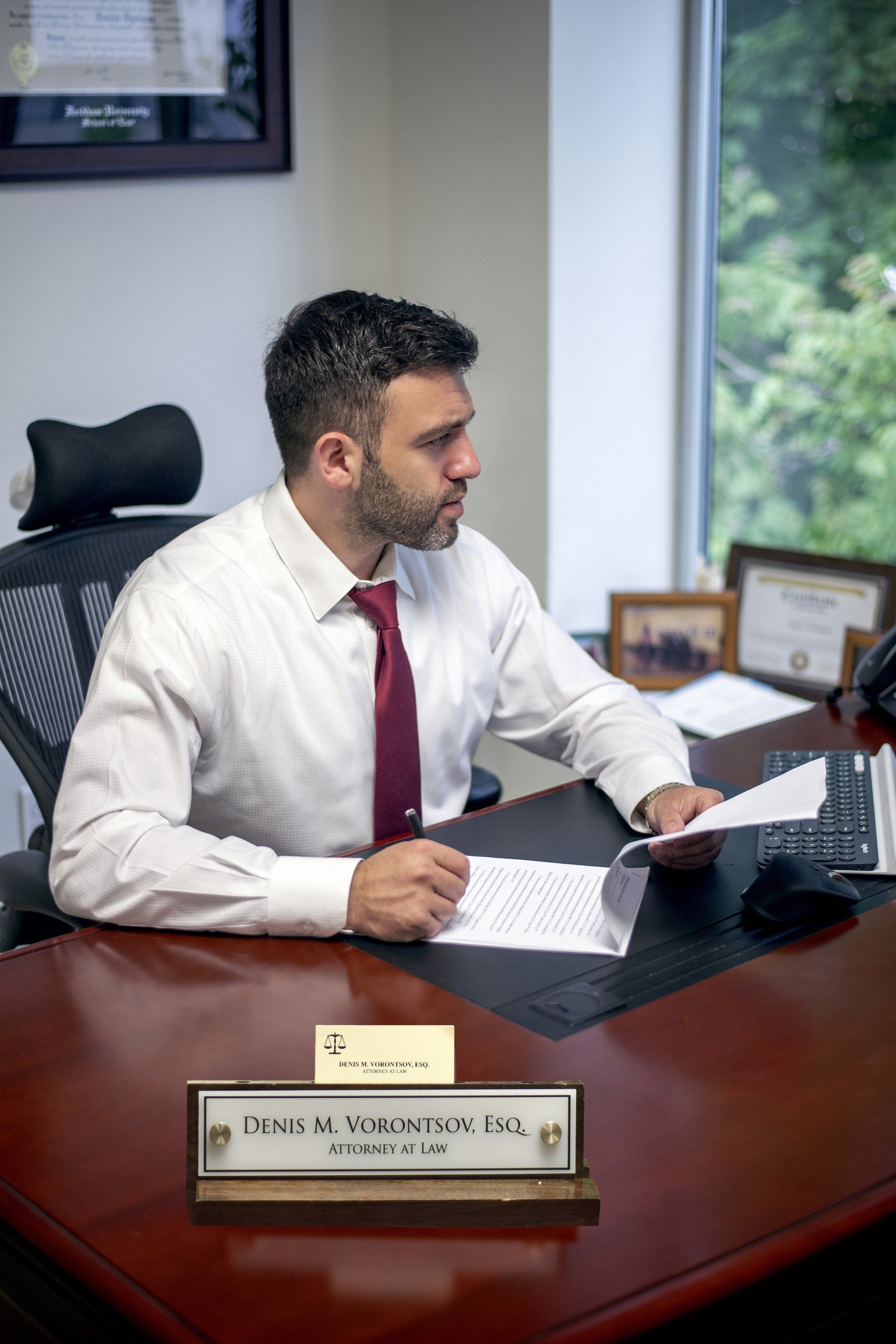 Man in a white shirt and red tie seated at an office desk, reading a document by a window.