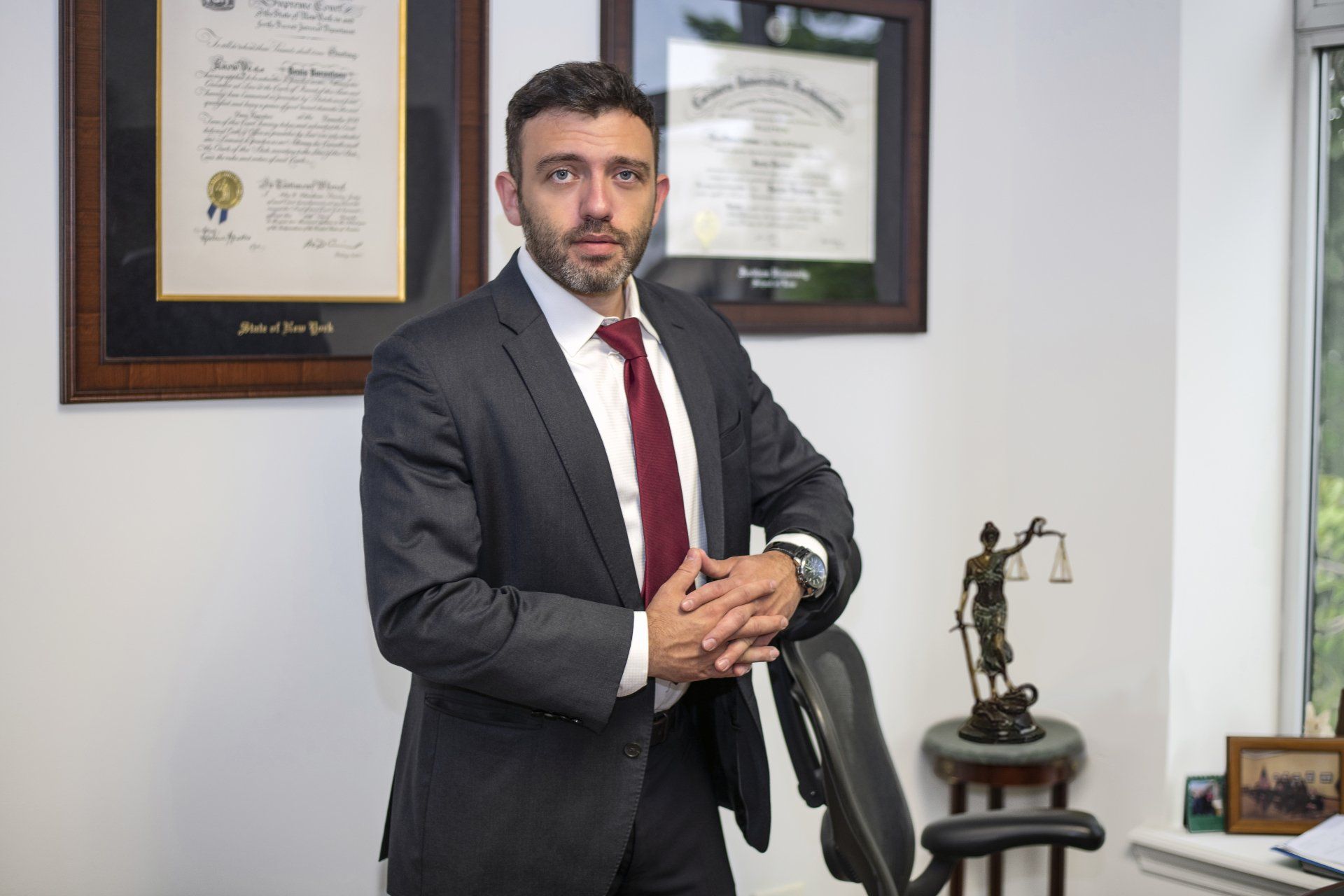 Man in a dark suit standing in an office beside framed certificates and a small statue