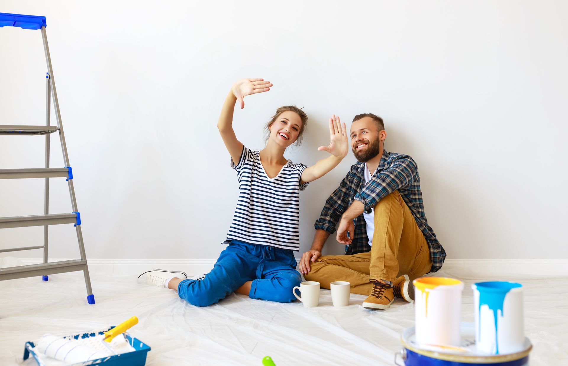 Two people painting a room, sitting on the floor with brushes, ladder and paint cans nearby