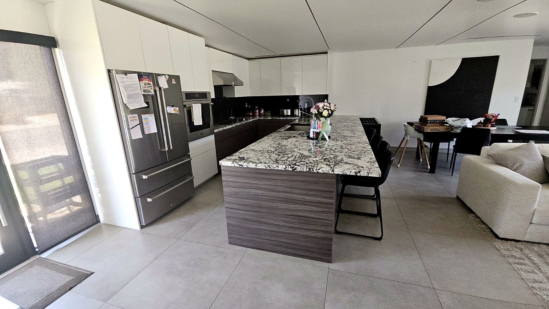 Modern open-plan kitchen and living room with granite island, stainless fridge, beige sofa, and tiled floor