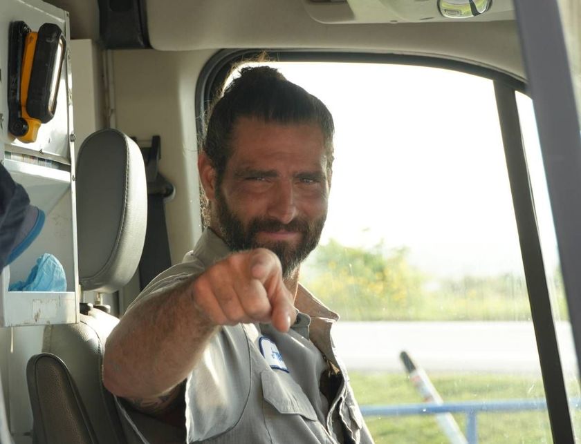 Smiling man in a vehicle points toward the camera, with a bright roadside visible outside the window.