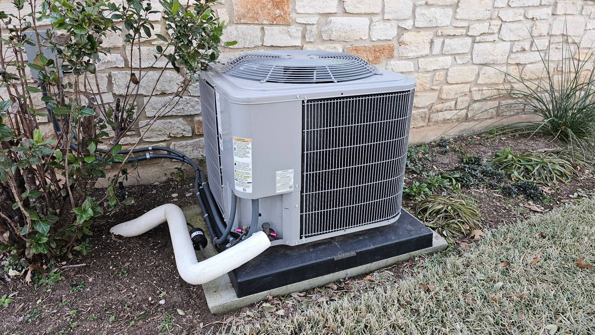 Outdoor HVAC unit beside a stone wall, with white pipes and a black grille on a gravel bed.