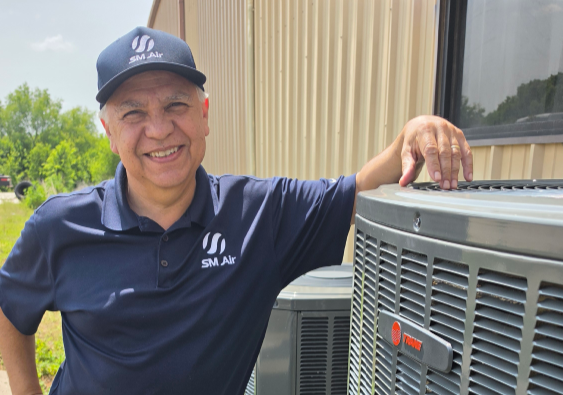 Smiling man in navy polo and cap leaning on an outdoor HVAC unit beside a building