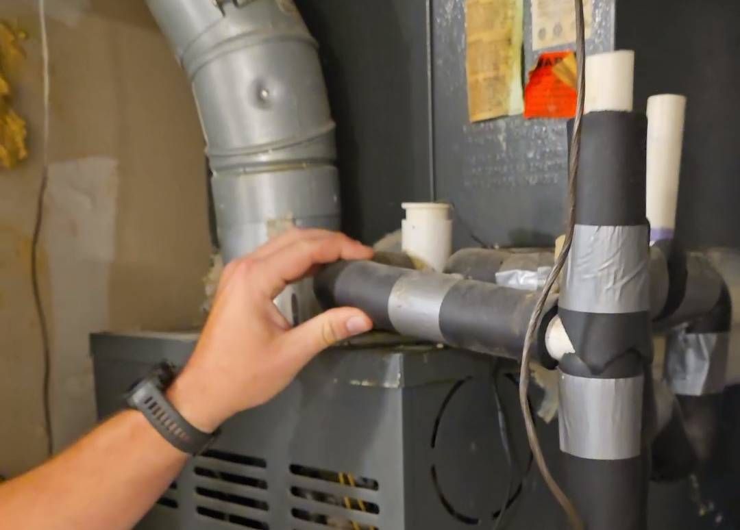 Hand holding a gray insulated pipe near a furnace with ductwork and wiring in a utility room.