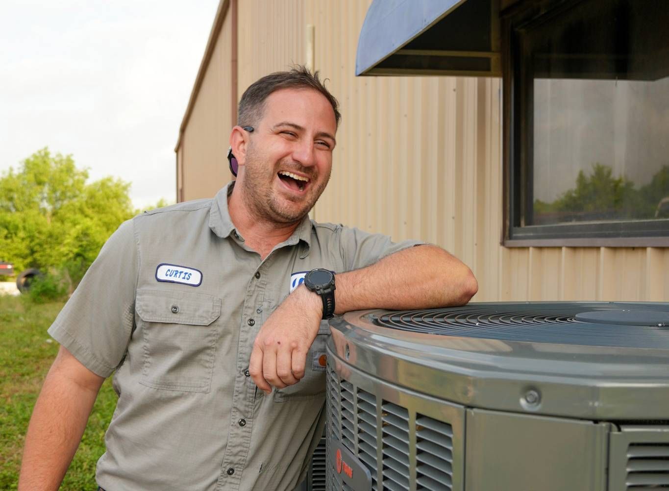 Man leaning on an outdoor HVAC unit beside a beige building, smiling and wearing a gray work shirt