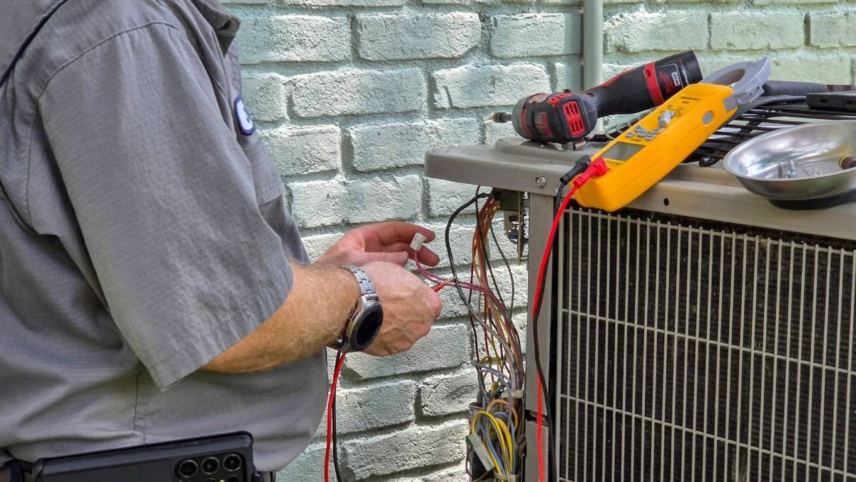Technician testing wiring on an outdoor HVAC unit with a multimeter and tools.
