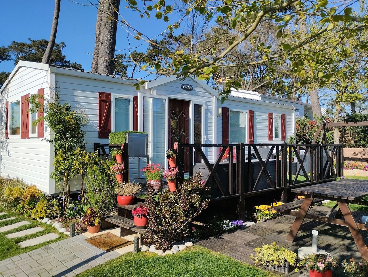 Small white mobile home with red shutters and a black porch railing in a sunny garden