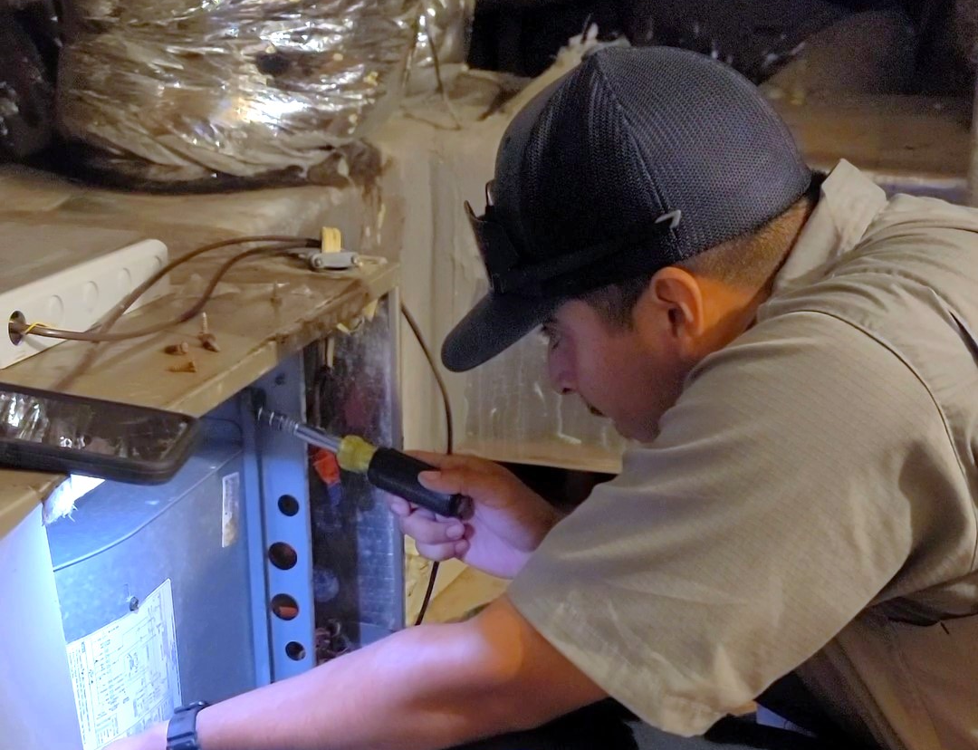 Technician inspecting an HVAC system with a flashlight and tool in an attic