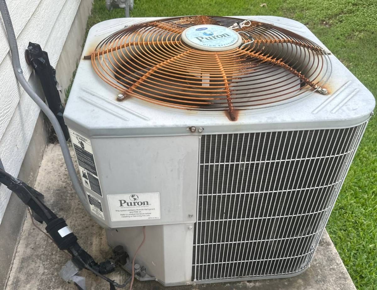 Outdoor air conditioner unit with a rust-orange fan grille and side vents, beside a wall on a concrete pa