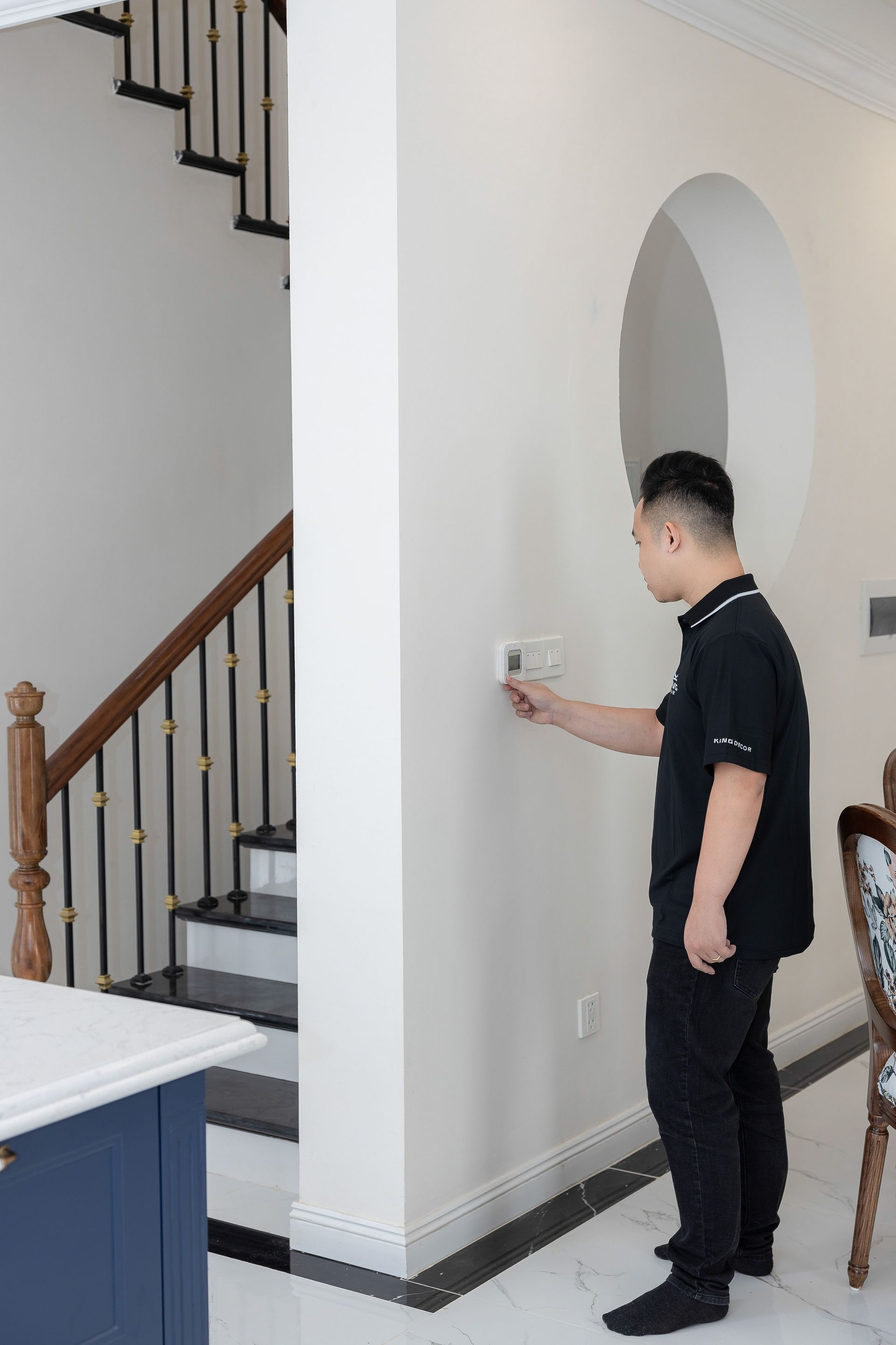 Man in black stands by a wall thermostat in a home hallway near a staircase.