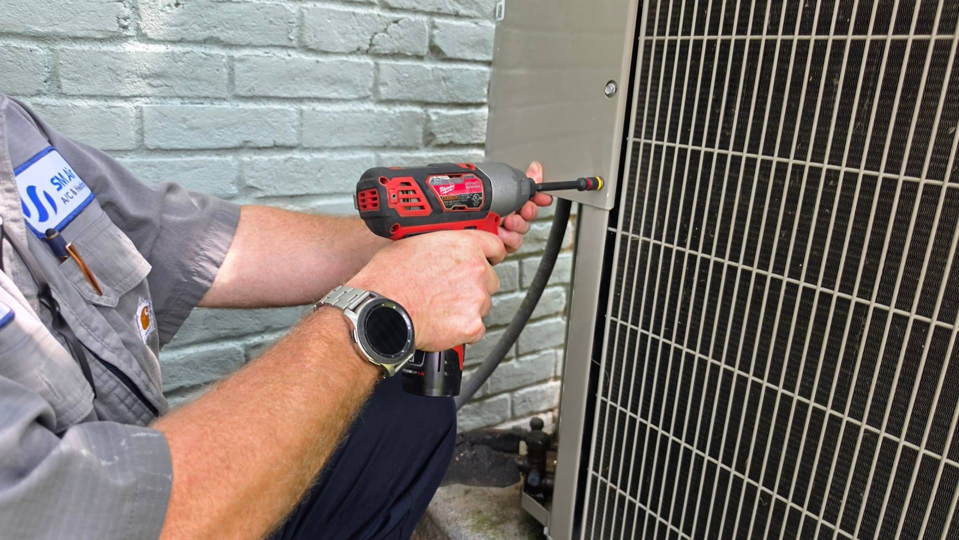 Worker using a red drill to fasten a metal vent grille to a brick wall