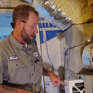 Technician in gray uniform points at a duct component in an unfinished basement with exposed HVAC ducts.