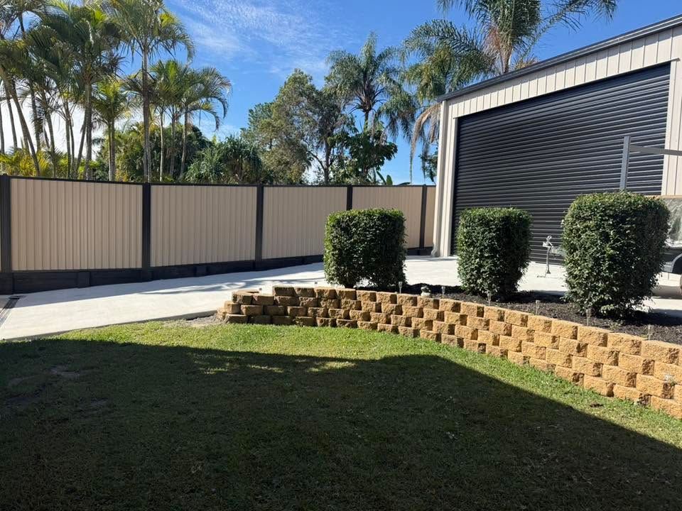 Landscaped backyard with green lawn, stone retaining wall, hedges, palm trees, and a modern garage