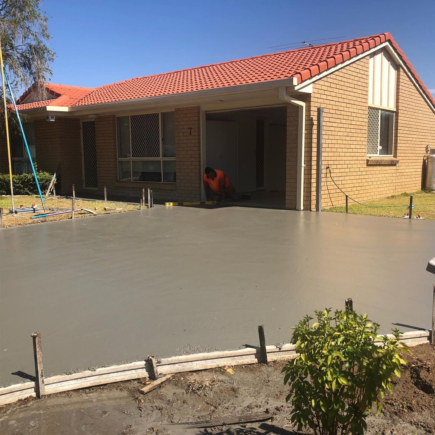 Single-story brick house with a red tile roof and freshly poured concrete driveway; worker under the carport
