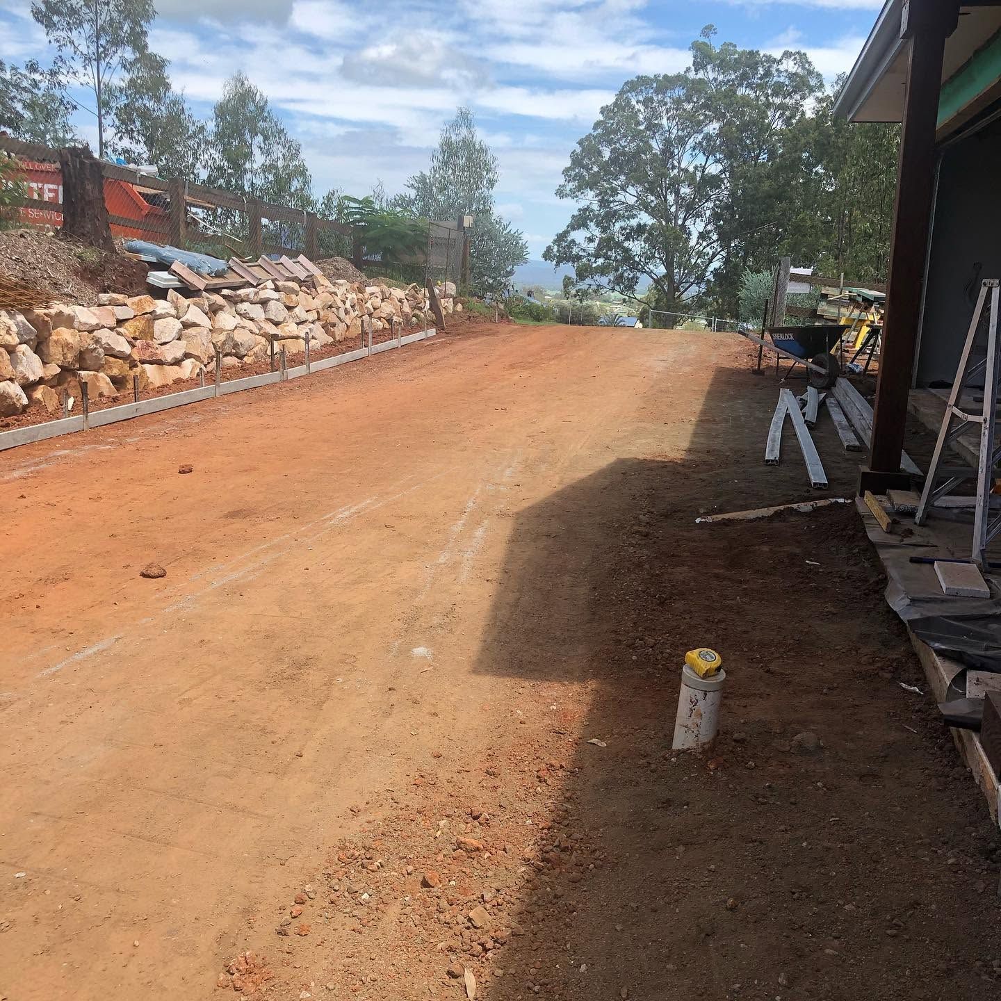 Dirt driveway under construction beside a stone retaining wall and trees, with tools on the right.