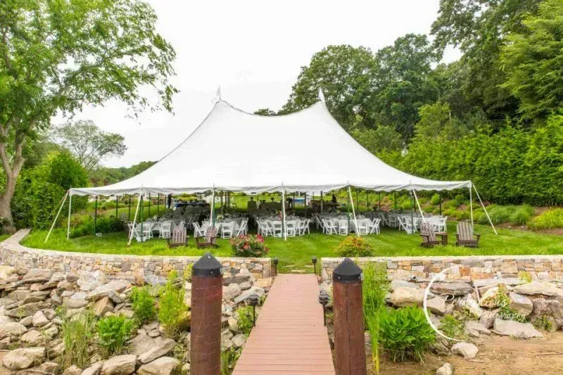 A large white marquee tent with dinner tables set up on a grassy lawn behind a stone retaining wall and a wooden dock.